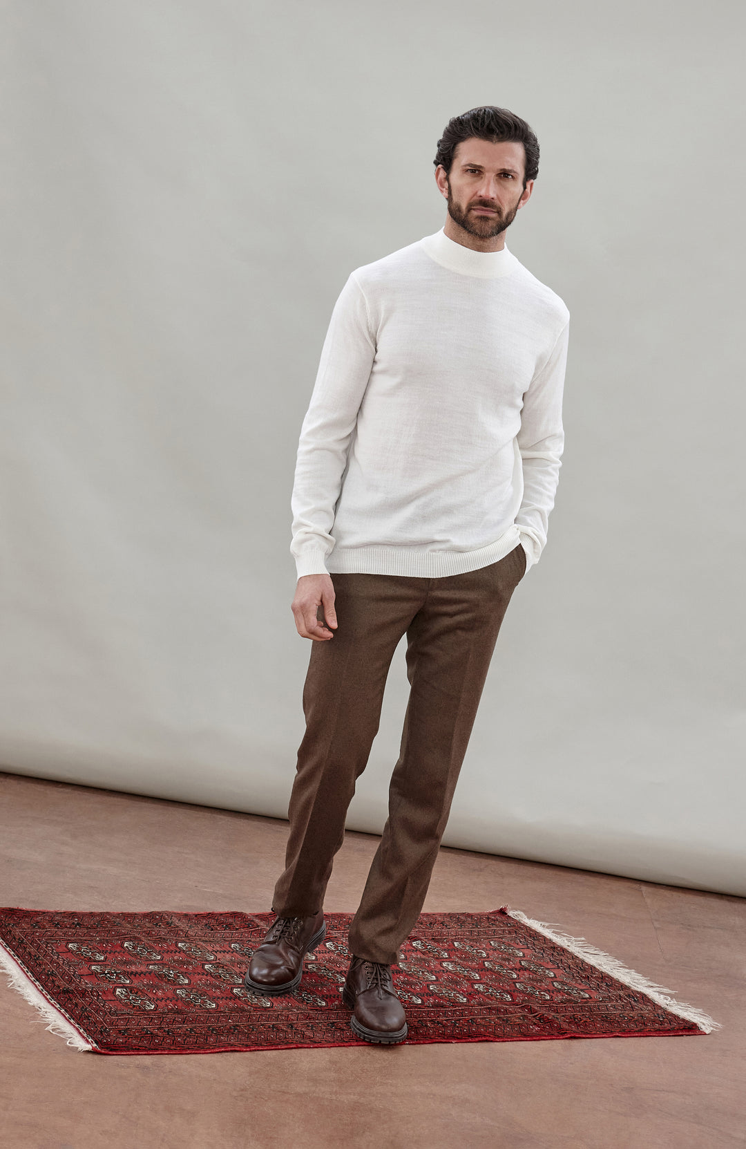 A man wearing a off white colored merino wool knit mock neck sweater and brown trousers, photographed against a neutral light gray studio background.