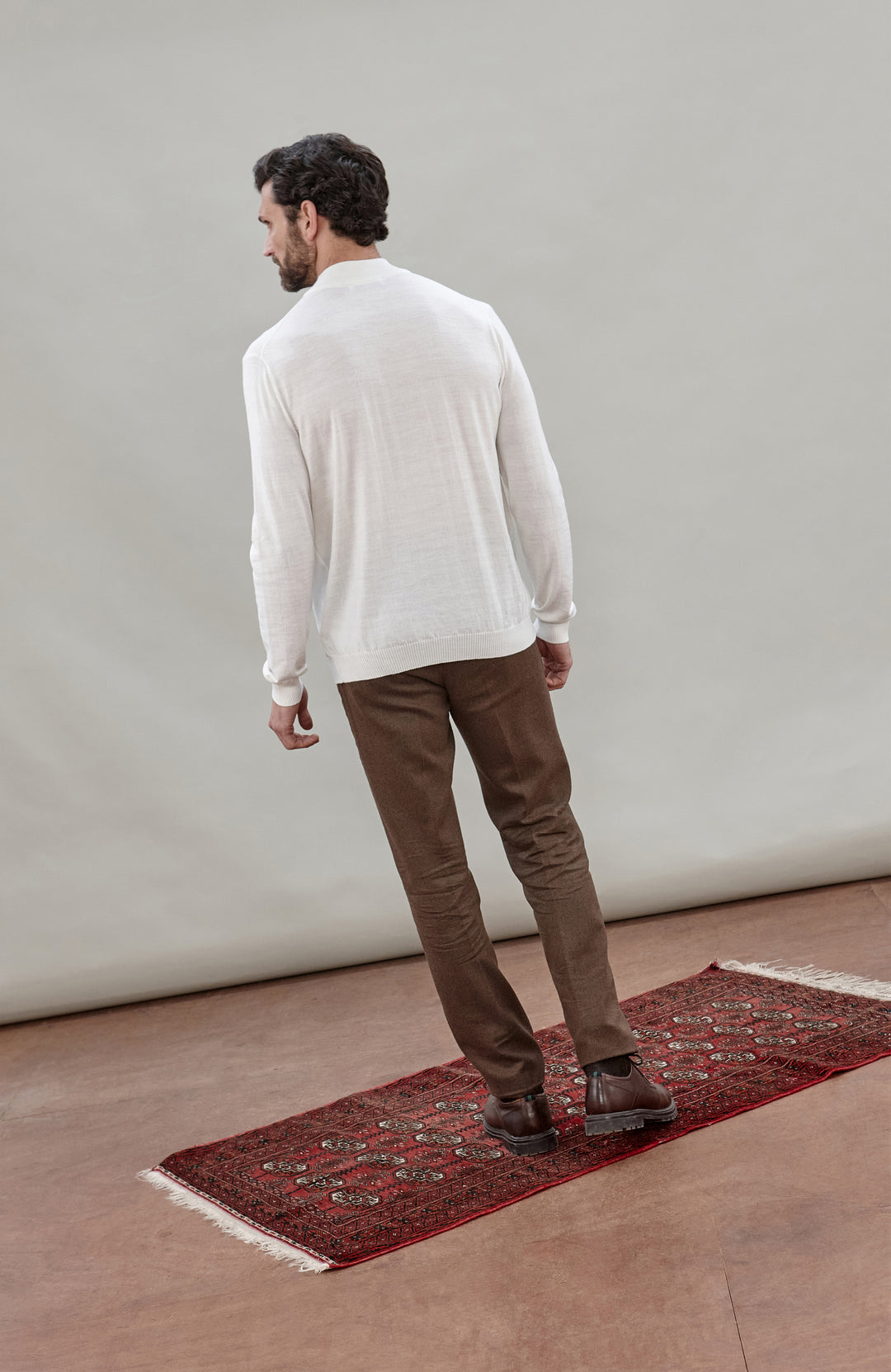 A man stands with his back to the camera wearing a white long-sleeve knit sweater, brown trousers, and brown shoes, standing on a red patterned runner rug against a neutral studio backdrop.