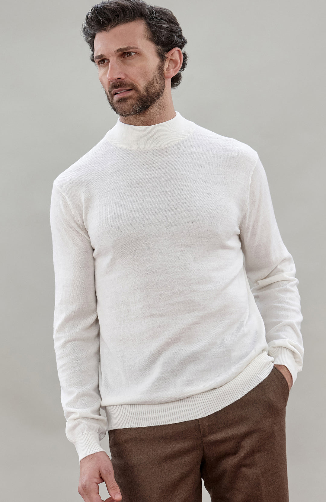 A man wearing a off white colored merino wool knit mock neck sweater and brown trousers, photographed against a neutral light gray studio background.