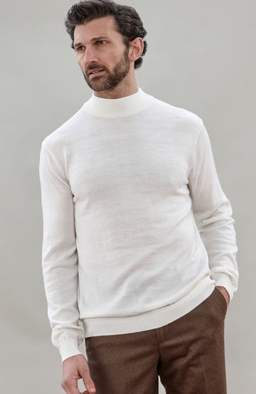 A man wearing a off white colored merino wool knit mock neck sweater and brown trousers, photographed against a neutral light gray studio background.