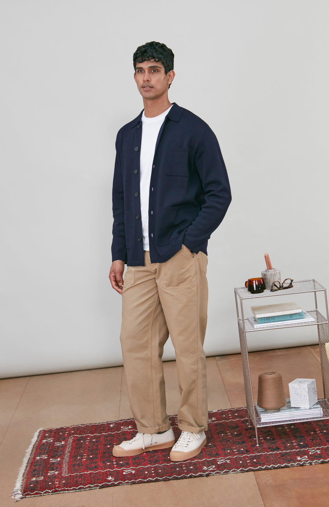 A man wearing a dark navy fine merino blue button-up shirt jacket by paul james knitwear with a collar and chest pocket, paired with beige trousers, photographed against a light gray studio background.