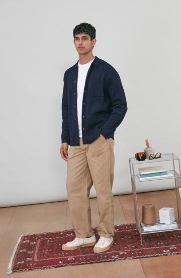 A man wearing a dark navy fine merino blue button-up shirt jacket by paul james knitwear with a collar and chest pocket, paired with beige trousers, photographed against a light gray studio background.