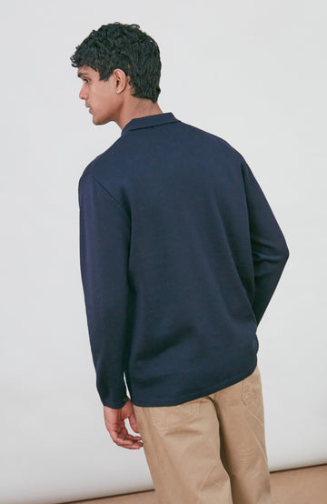 A man wearing a dark navy fine merino blue button-up shirt jacket by paul james knitwear with a collar and chest pocket, paired with beige trousers, photographed against a light gray studio background.