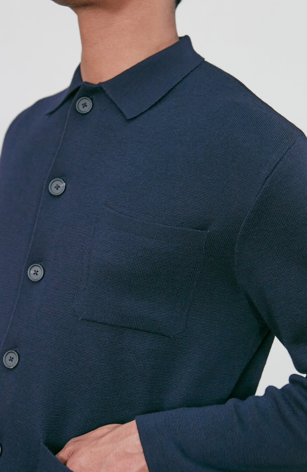 A man wearing a dark navy fine merino blue button-up shirt jacket by paul james knitwear with a collar and chest pocket, paired with beige trousers, photographed against a light gray studio background.