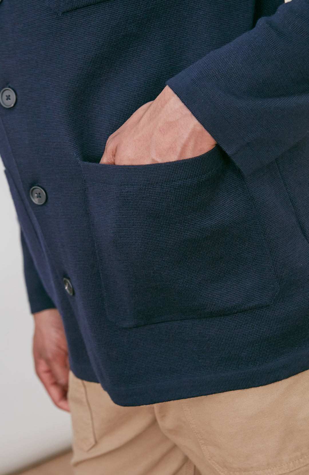 A man wearing a dark navy fine merino blue button-up shirt jacket by paul james knitwear with a collar and chest pocket, paired with beige trousers, photographed against a light gray studio background.