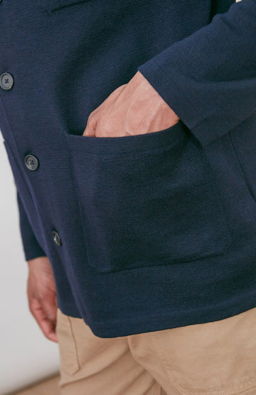 A man wearing a dark navy fine merino blue button-up shirt jacket by paul james knitwear with a collar and chest pocket, paired with beige trousers, photographed against a light gray studio background.