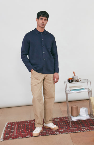 A man wearing a dark navy fine merino blue button-up shirt jacket by paul james knitwear with a collar and chest pocket, paired with beige trousers, photographed against a light gray studio background.