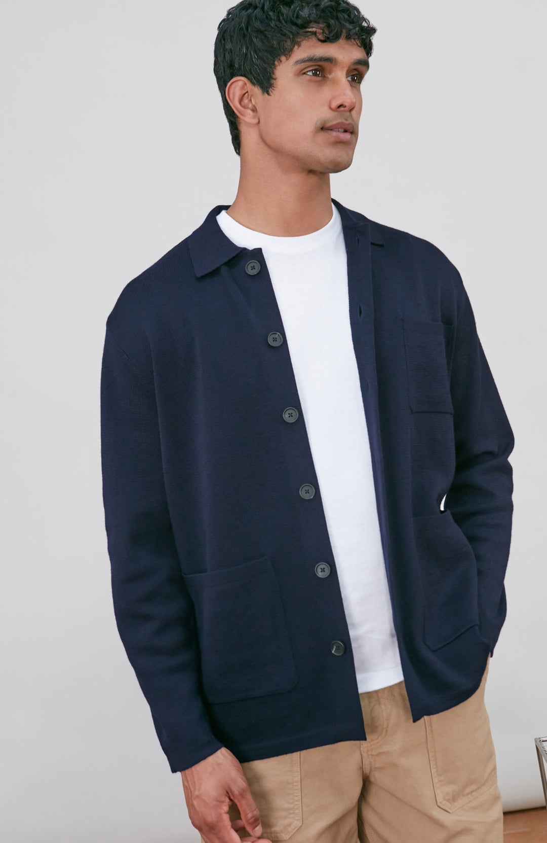 A man wearing a dark navy fine merino blue button-up shirt jacket by paul james knitwear with a collar and chest pocket, paired with beige trousers, photographed against a light gray studio background.