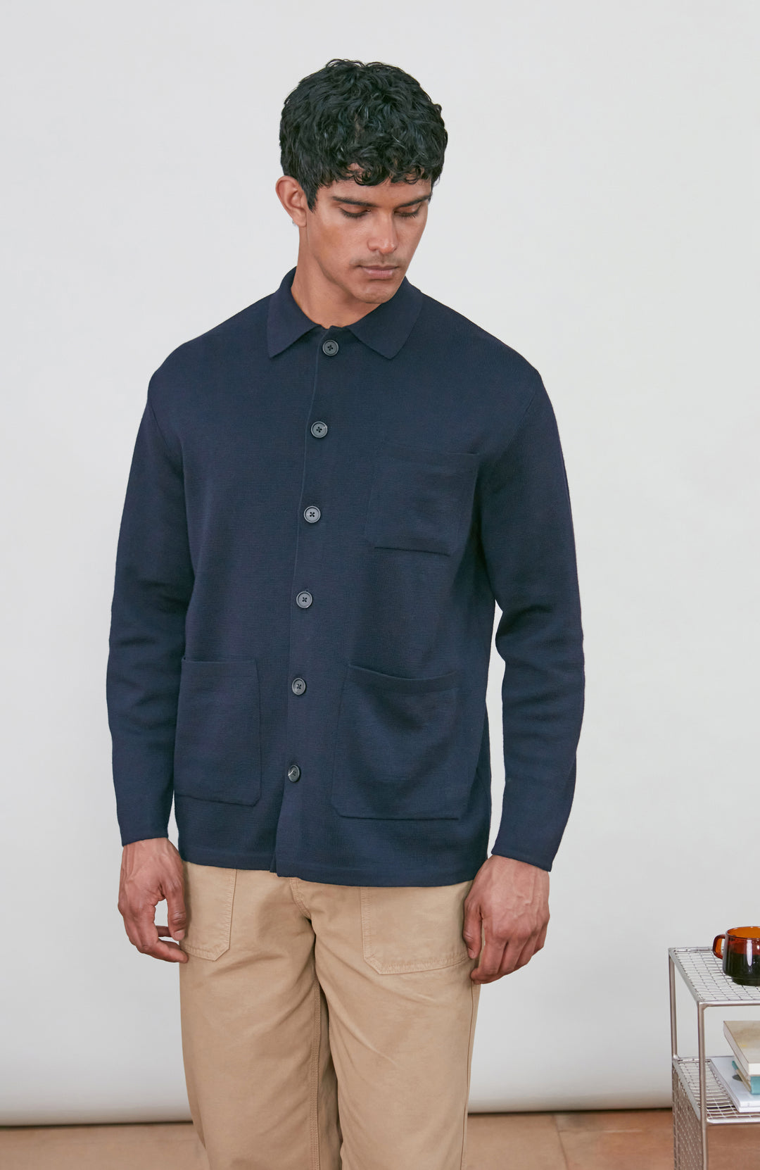 A man wearing a dark navy fine merino blue button-up shirt jacket by paul james knitwear with a collar and chest pocket, paired with beige trousers, photographed against a light gray studio background.