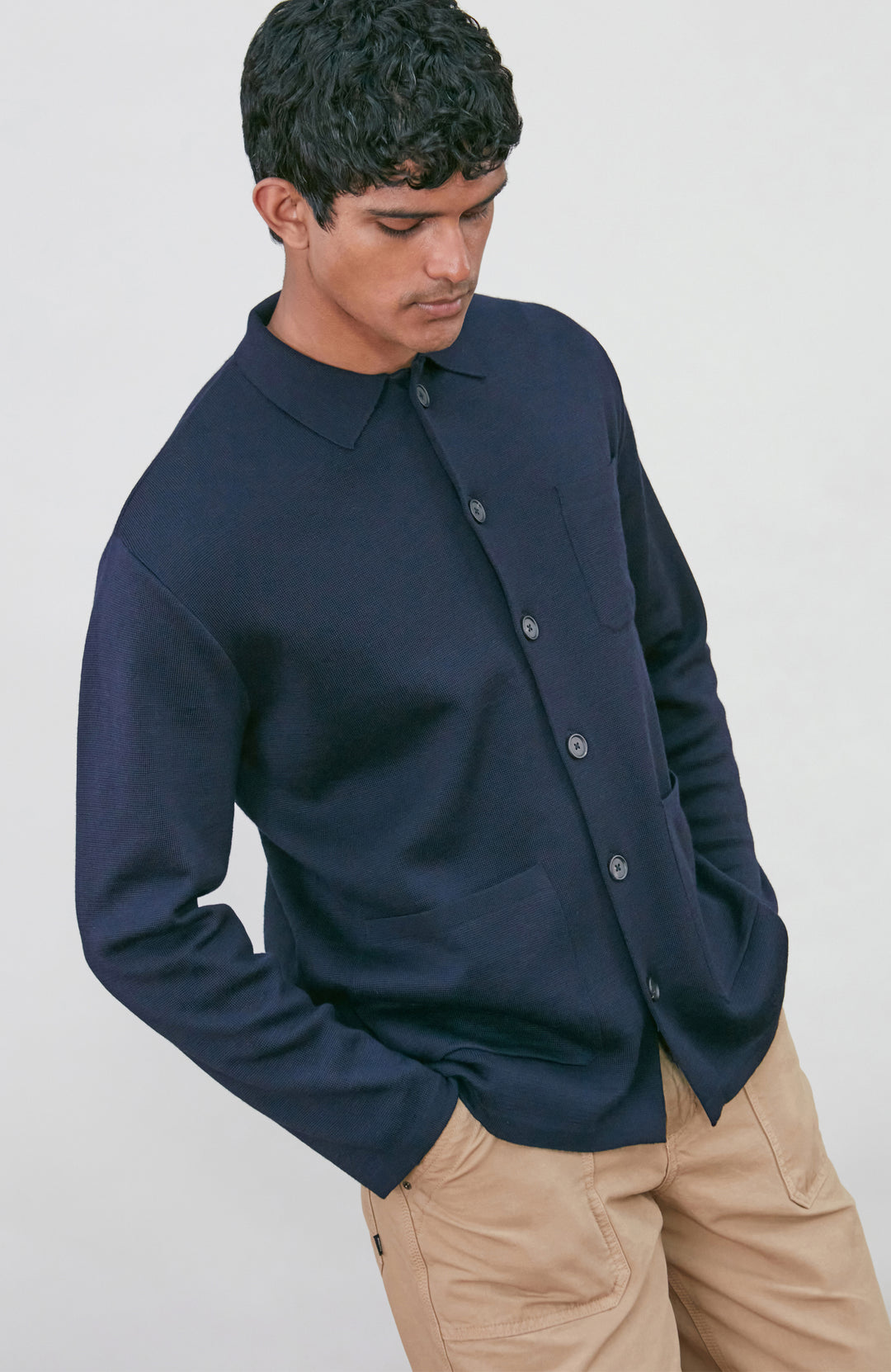 A man wearing a dark navy fine merino blue button-up shirt jacket by paul james knitwear with a collar and chest pocket, paired with beige trousers, photographed against a light gray studio background.