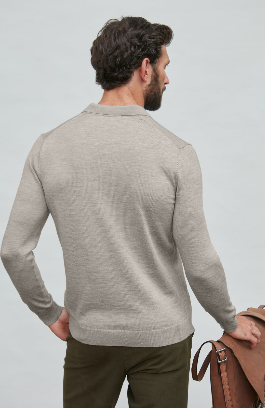 Man wearing a gray sweater and holding a brown leather bag against a light gray background by Paul James
