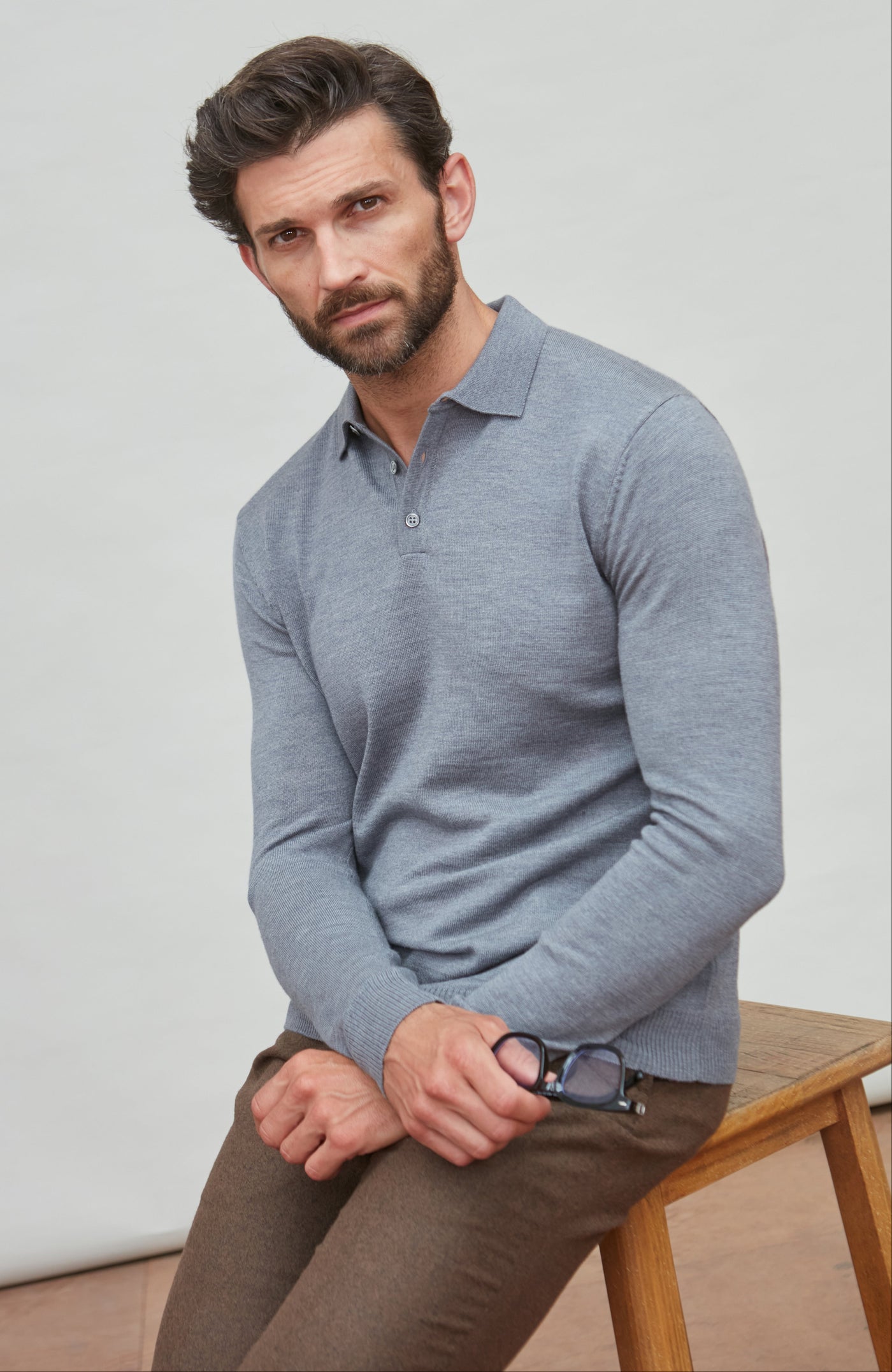 Man wearing a light grey polo neck merino wool sweater sitting on a wooden bench against a plain background