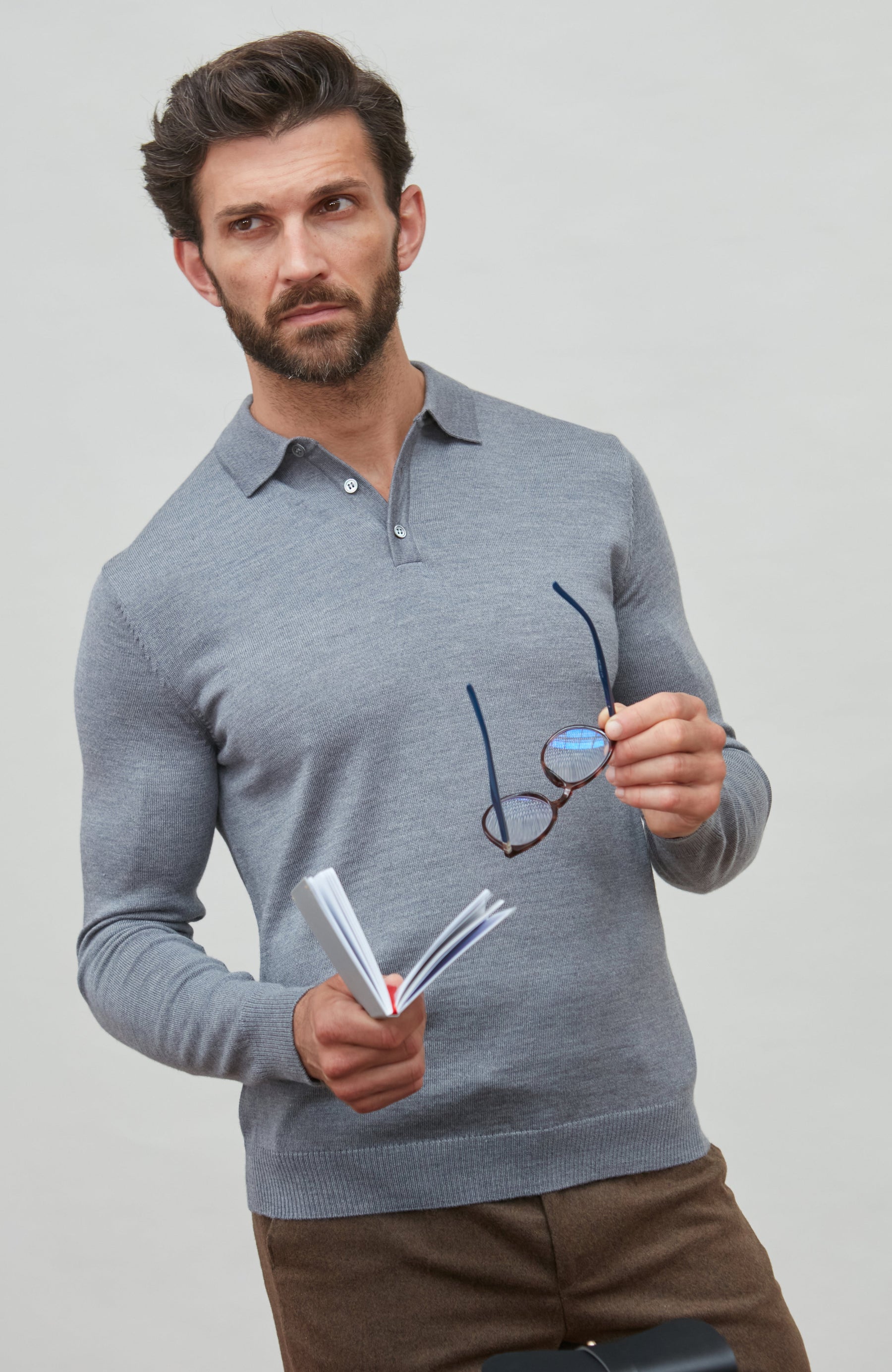 Man wearing a  light grey polo neck merino wool  sweater holding eyeglasses and a notebook against a light gray background by Paul James 