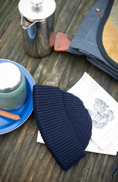 Navy wool fisherman knit beanie on a wooden surface with a teapot and saucer in the background