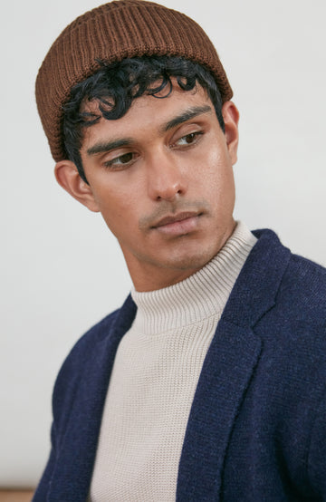 A man wearing a brown ribbed fine wool fisherman knit beanie by paul james knitwear, a cream ribbed turtleneck sweater, and a navy blue knit cardigan, photographed against a plain light gray background.