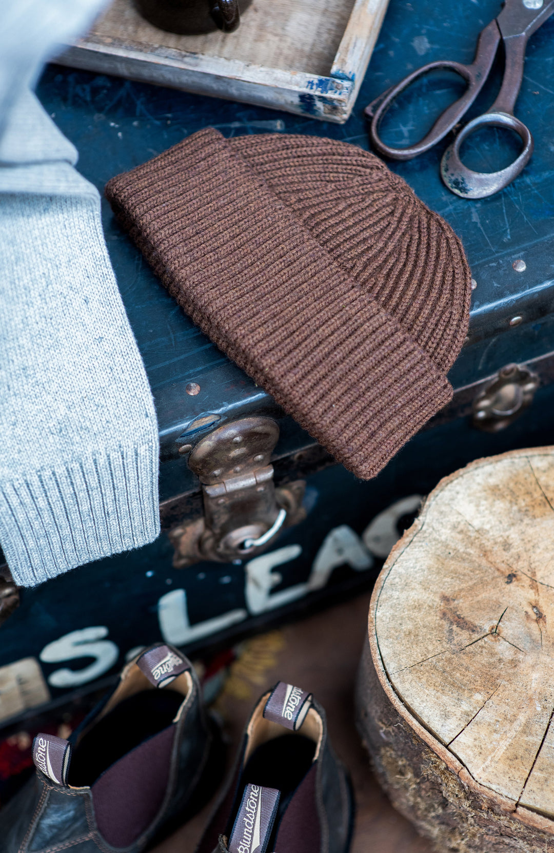 Brown knitted fisherman beanie on a vintage suitcase with sunglasses and a pair of scissors in the background.