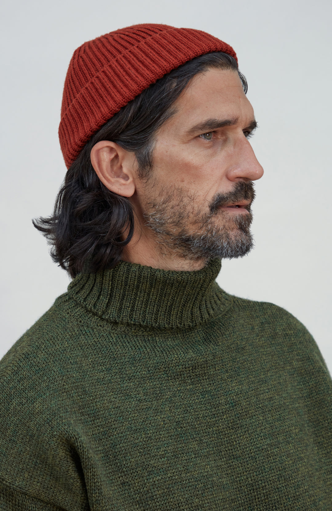A man in profile wearing a rust-red ribbed knit beanie and an olive green ribbed knit turtleneck sweater, photographed against a plain light gray background. Fine merino wool fisherman beanie hat by Paul James Knitwear.