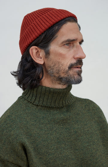 A man in profile wearing a rust-red ribbed knit beanie and an olive green ribbed knit turtleneck sweater, photographed against a plain light gray background. Fine merino wool fisherman beanie hat by Paul James Knitwear.