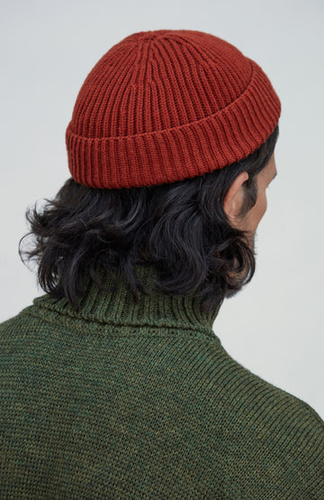 A person shown from the back wearing a rust-red ribbed knit beanie and an olive green knit turtleneck sweater against a light gray background. Fine merino wool fisherman beanie hat by Paul James Knitwear.