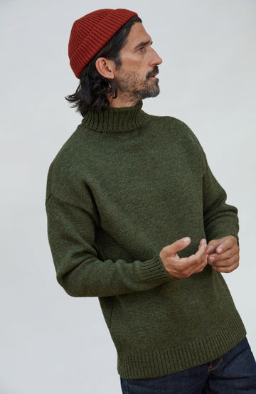 A man wearing an olive green knit turtleneck sweater, a rust red beanie, and dark blue jeans, photographed against a plain light gray studio background. Fine merino wool fisherman beanie hat by Paul James Knitwear.