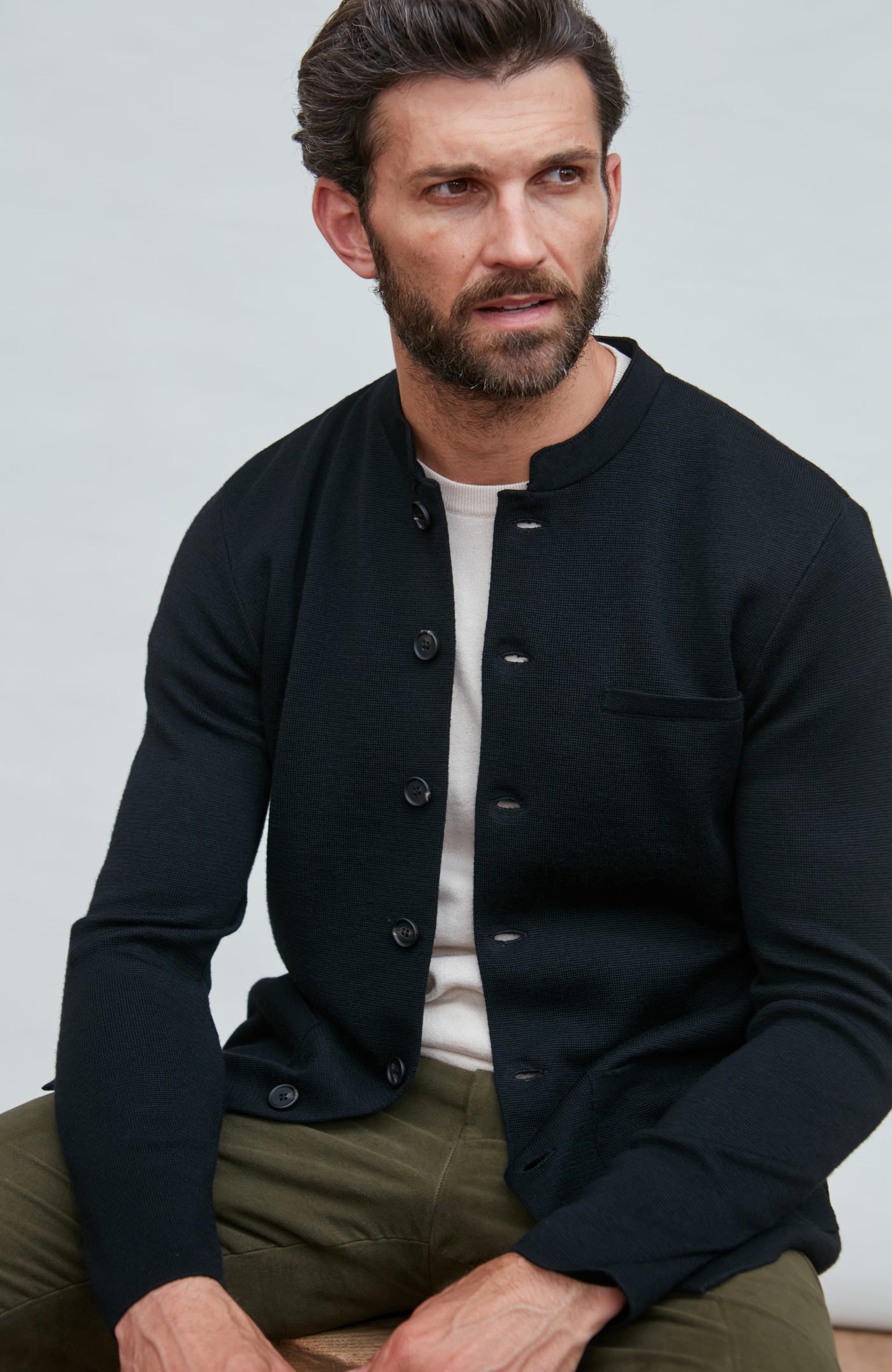 Man wearing a black mandarin collar jacket in fine wool over a white shirt and green pants against a light gray background