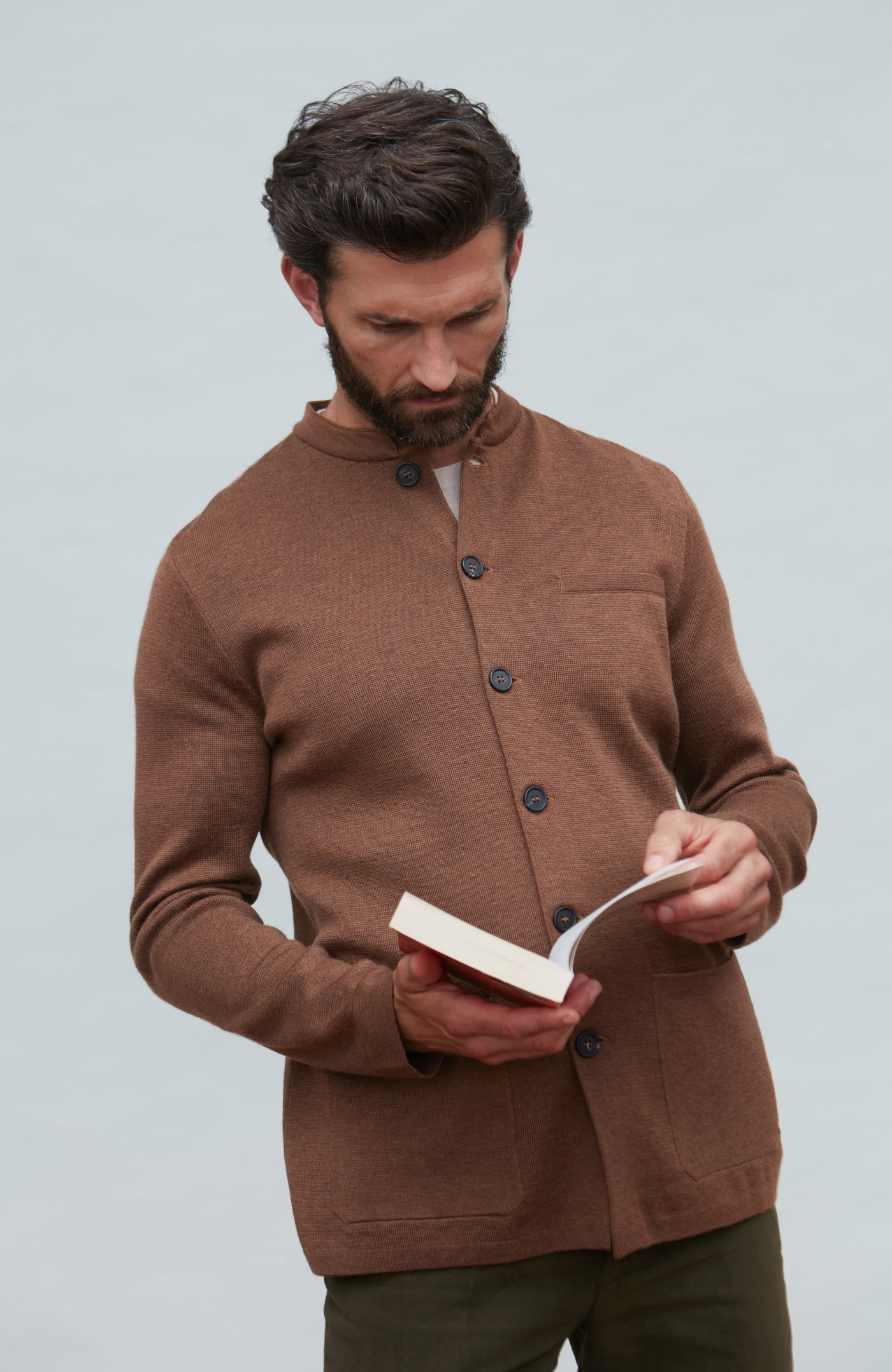 Man wearing a brown mandarin collar jacket in fine wool holding a book against a light gray background