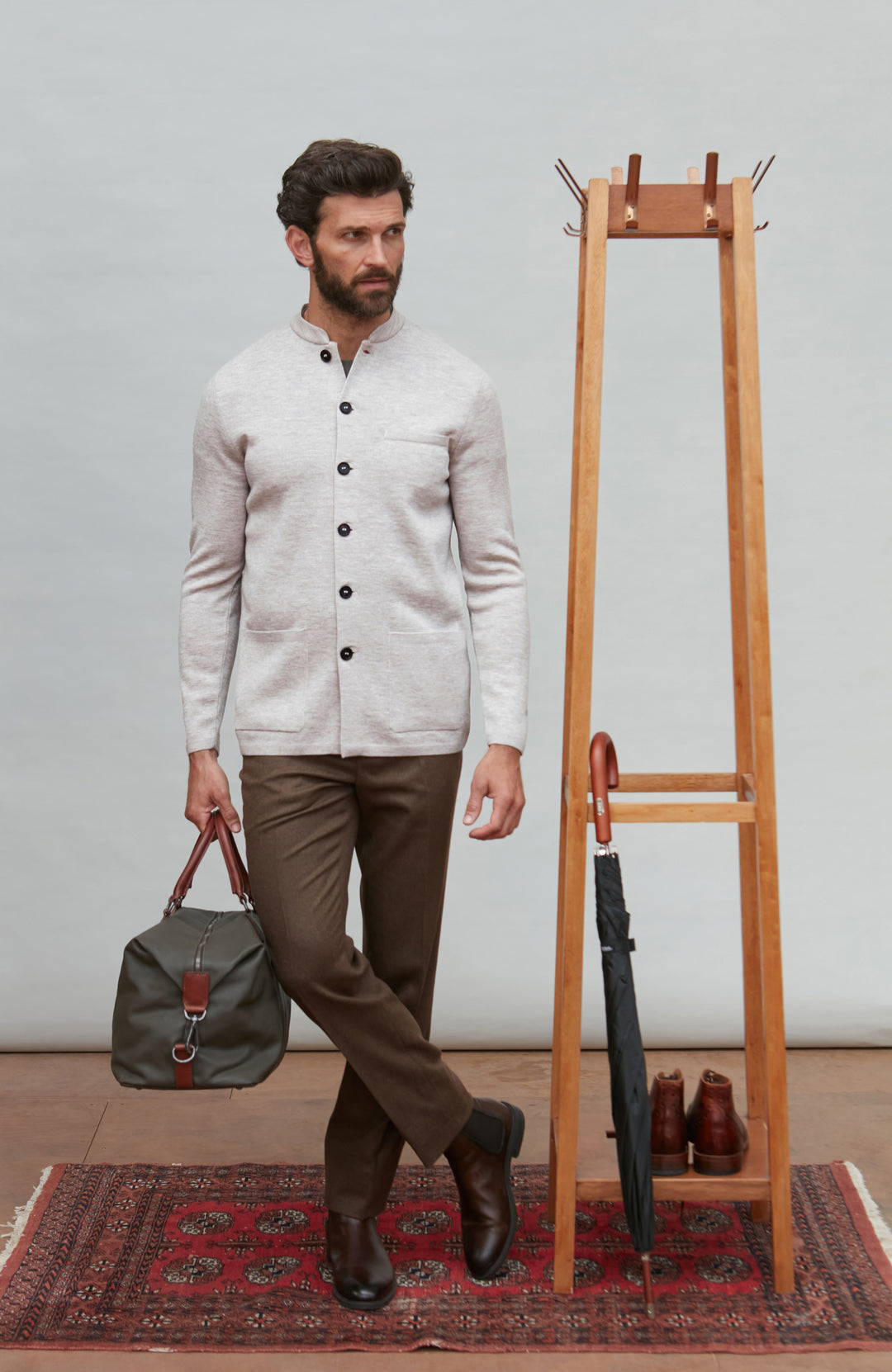 Man in a light-colored light gray mandarin collar jacket and brown pants holding a green bag, standing next to a wooden coat rack with shoes on a patterned rug by Paul James