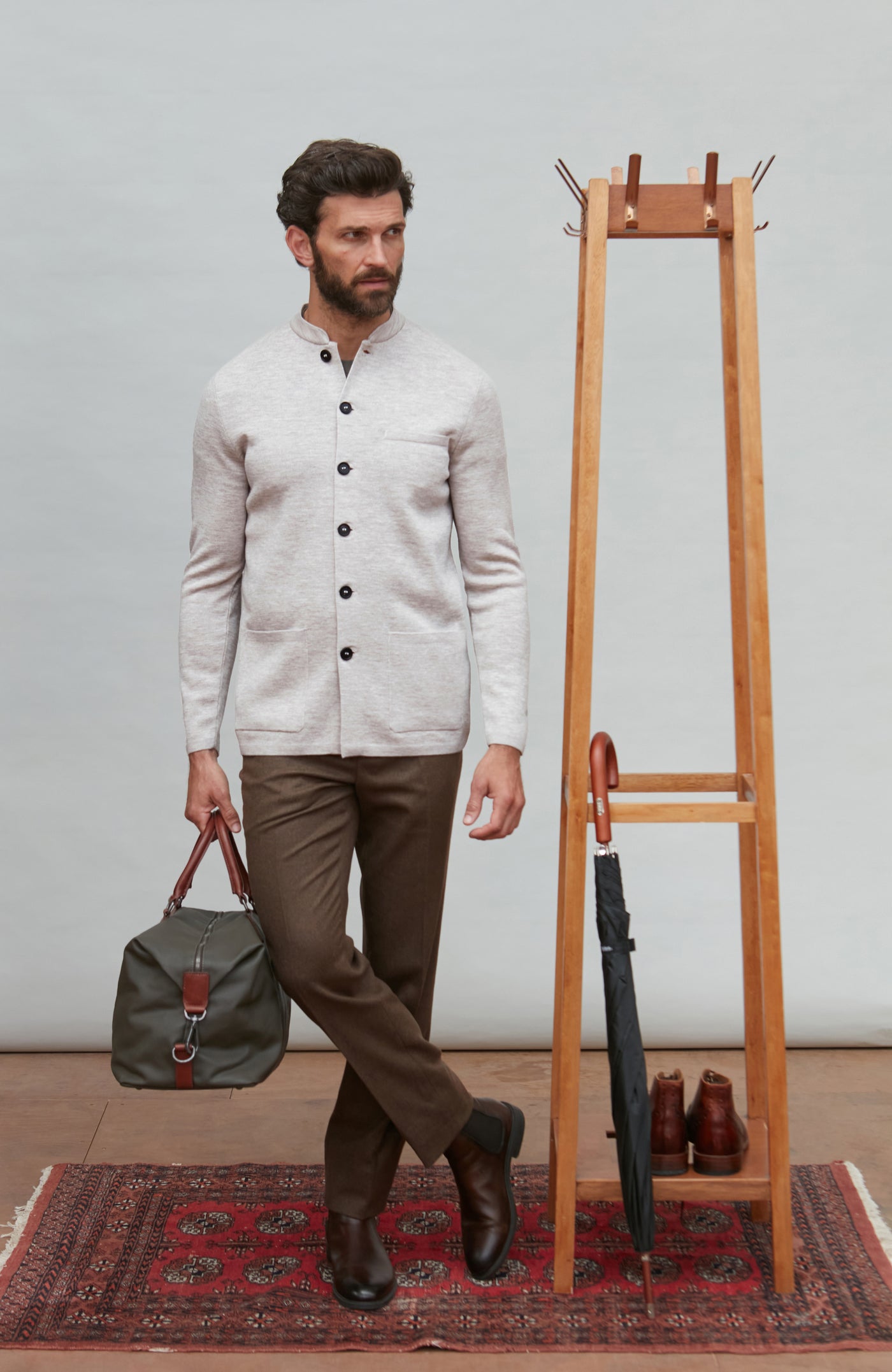 Man in a light-colored light gray mandarin collar jacket and brown pants holding a green bag, standing next to a wooden coat rack with shoes on a patterned rug.