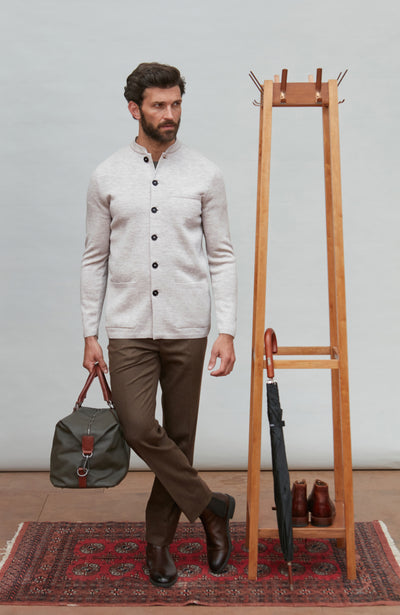 Man in a light-colored light gray mandarin collar jacket and brown pants holding a green bag, standing next to a wooden coat rack with shoes on a patterned rug.