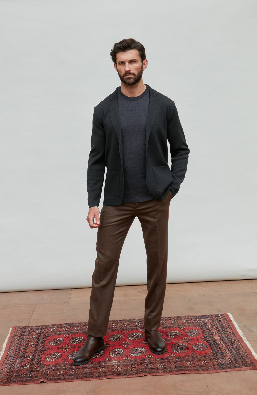 Man wearing a black fine merino wool and brown pants standing on a red patterned rug with a white wall background.
