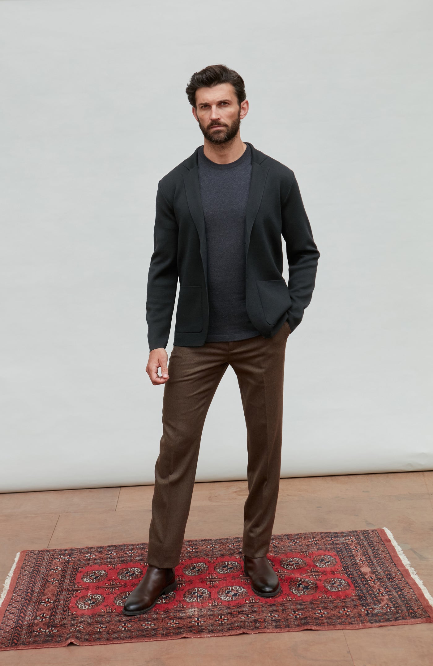 Man wearing a black fine merino wool and brown pants standing on a red patterned rug with a white wall background.