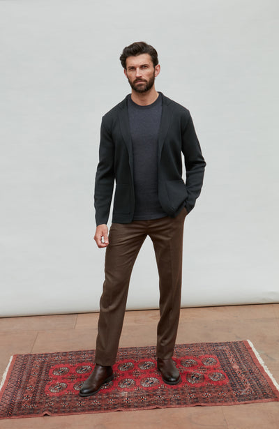 Man wearing a black fine merino wool and brown pants standing on a red patterned rug with a white wall background.