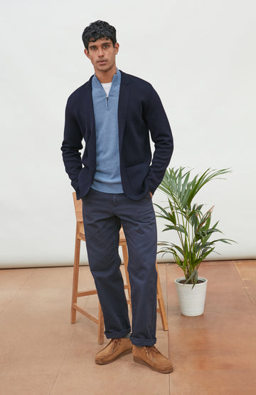 A man wearing a navy blue merino wool knit blazer by paul james knitwear over a light blue half-zip sweater and a white undershirt, standing against a plain light background with a plant and wooden chair partially visible.