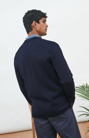 A man wearing a navy blue merino wool knit blazer by paul james knitwear over a light blue half-zip sweater and a white undershirt, standing against a plain light background with a plant and wooden chair partially visible.