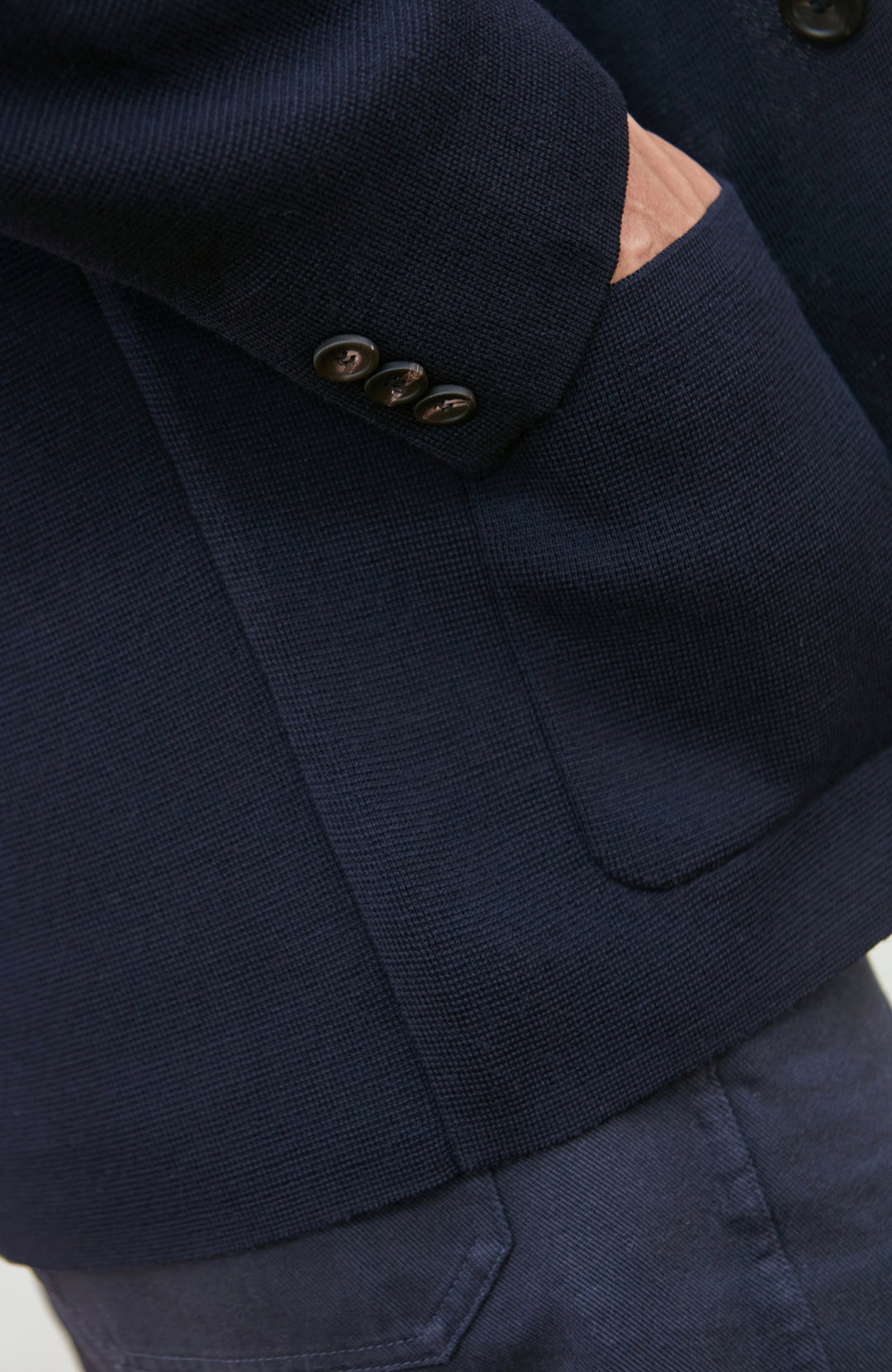 A man wearing a navy blue merino wool knit blazer by paul james knitwear over a light blue half-zip sweater and a white undershirt, standing against a plain light background with a plant and wooden chair partially visible.
