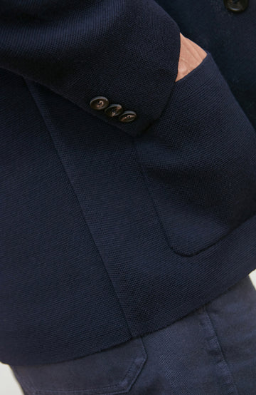 A man wearing a navy blue merino wool knit blazer by paul james knitwear over a light blue half-zip sweater and a white undershirt, standing against a plain light background with a plant and wooden chair partially visible.