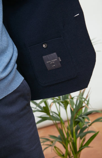 A man wearing a navy blue merino wool knit blazer by paul james knitwear over a light blue half-zip sweater and a white undershirt, standing against a plain light background with a plant and wooden chair partially visible.