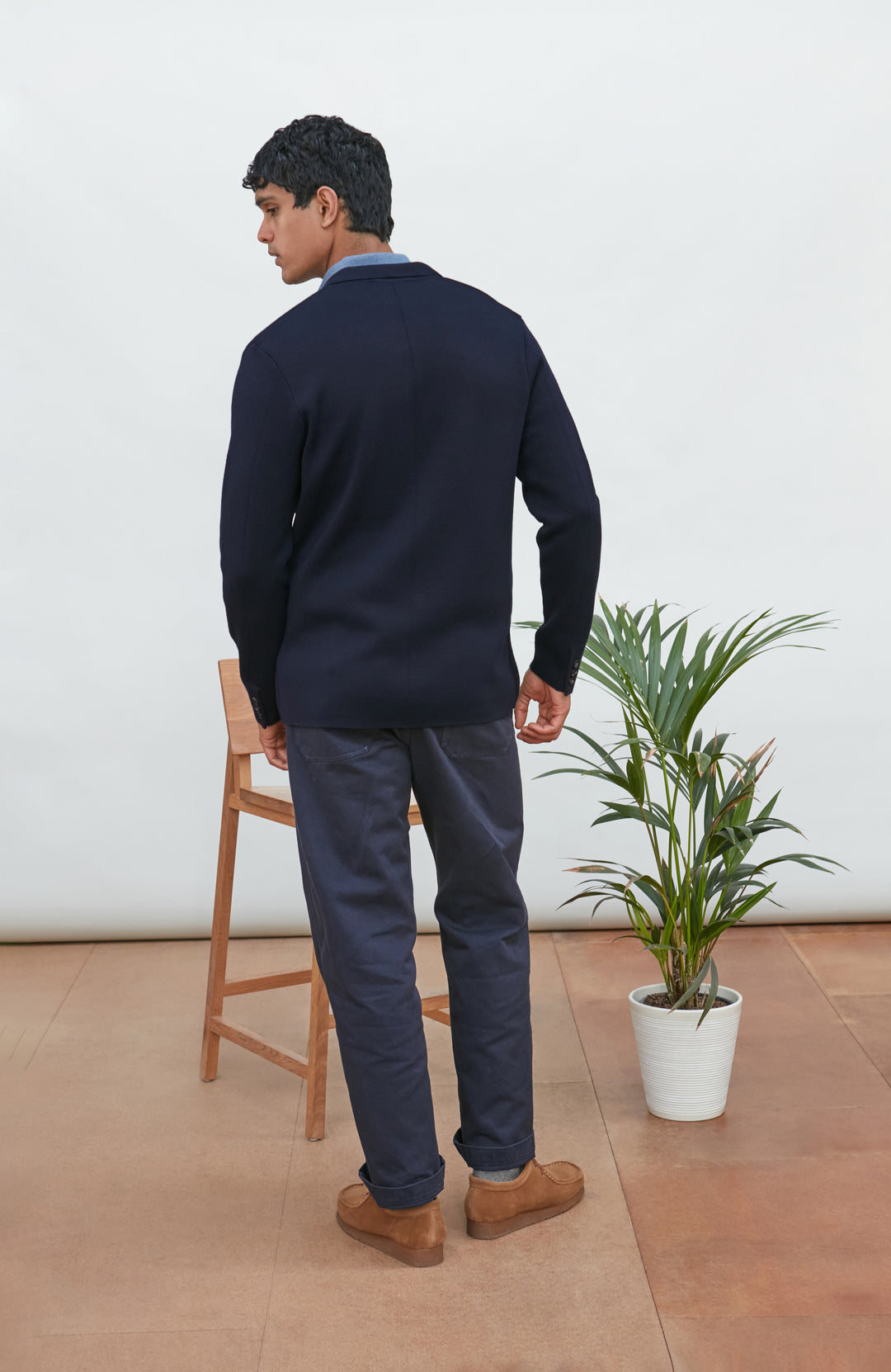 A man wearing a navy blue merino wool knit blazer by paul james knitwear over a light blue half-zip sweater and a white undershirt, standing against a plain light background with a plant and wooden chair partially visible.