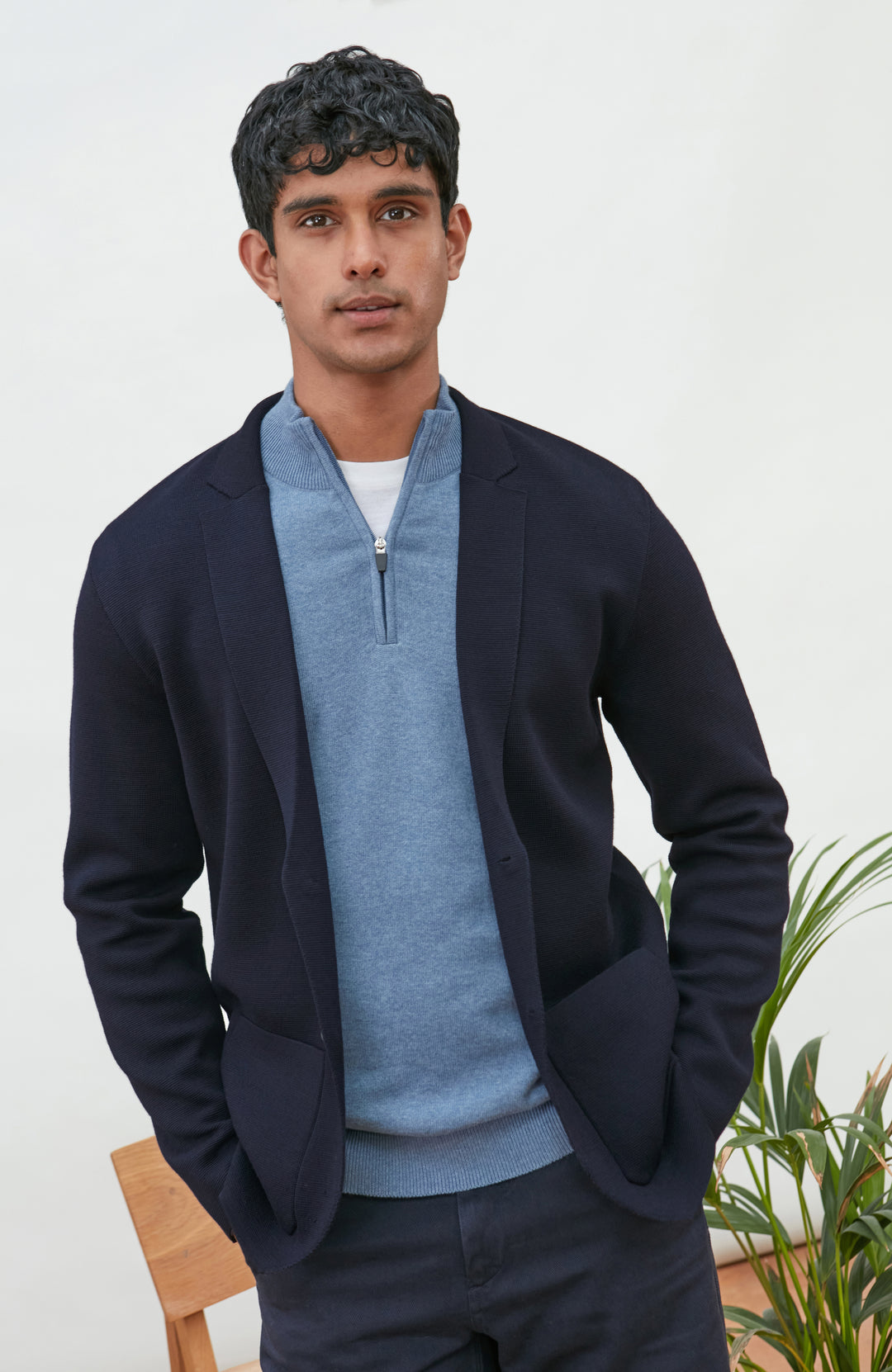 A man wearing a navy blue merino wool knit blazer by paul james knitwear over a light blue half-zip sweater and a white undershirt, standing against a plain light background with a plant and wooden chair partially visible.