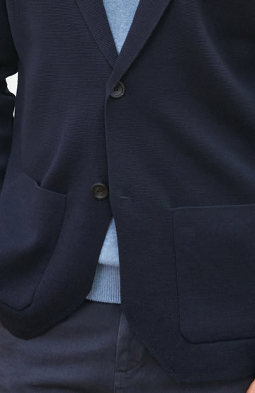 A man wearing a navy blue merino wool knit blazer by paul james knitwear over a light blue half-zip sweater and a white undershirt, standing against a plain light background with a plant and wooden chair partially visible.