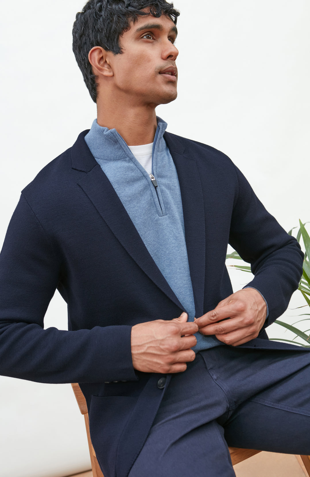 A man wearing a navy blue merino wool knit blazer by paul james knitwear over a light blue half-zip sweater and a white undershirt, standing against a plain light background with a plant and wooden chair partially visible.