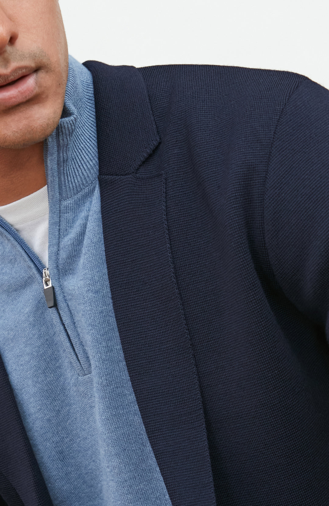 A man wearing a navy blue merino wool knit blazer by paul james knitwear over a light blue half-zip sweater and a white undershirt, standing against a plain light background with a plant and wooden chair partially visible.
