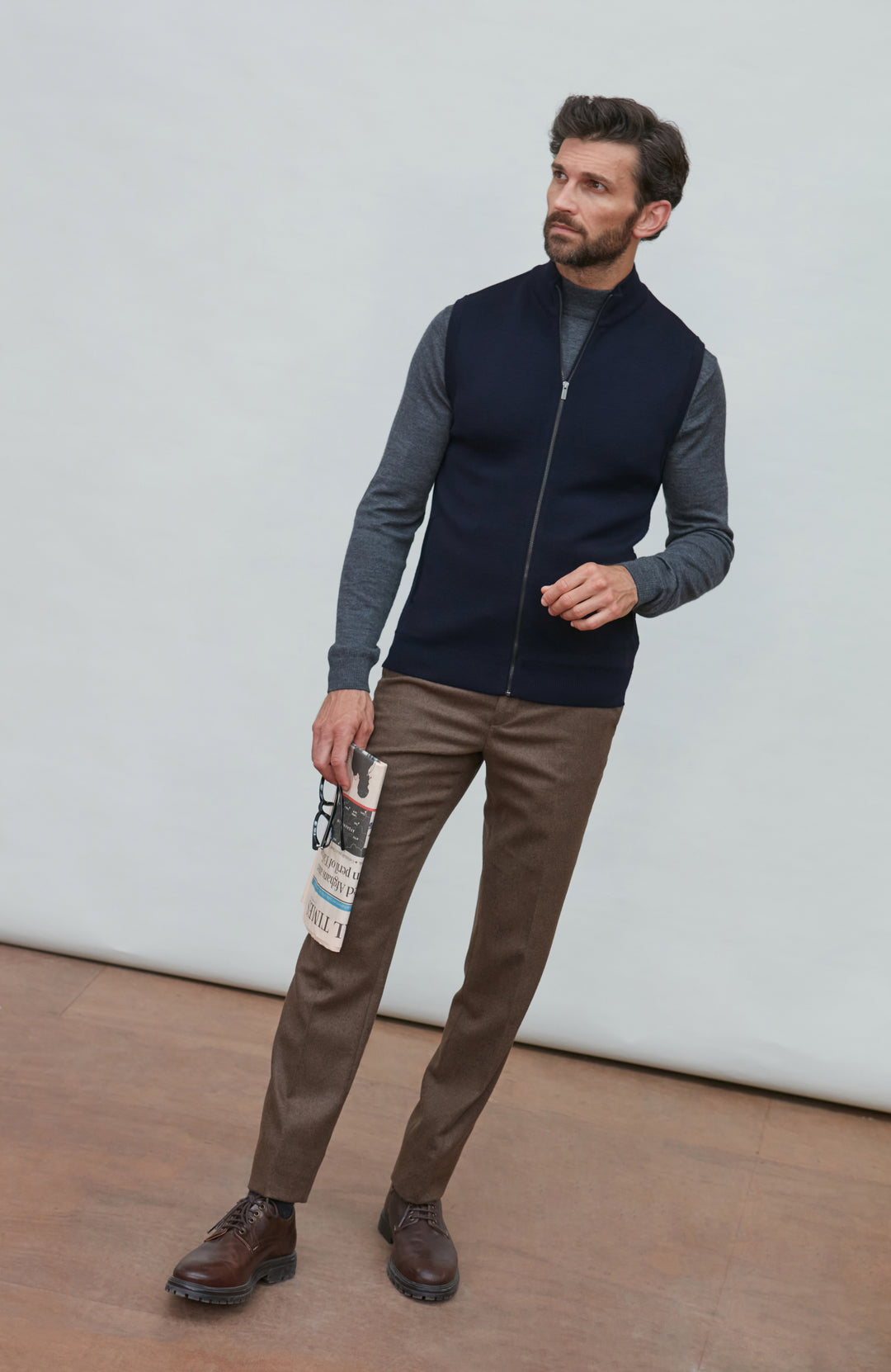 A man wearing a navy blue zip-up sleeveless merino wool vest by Paul James Knitwear over a gray long-sleeve shirt, brown trousers, and brown leather shoes, standing against a light gray wall.