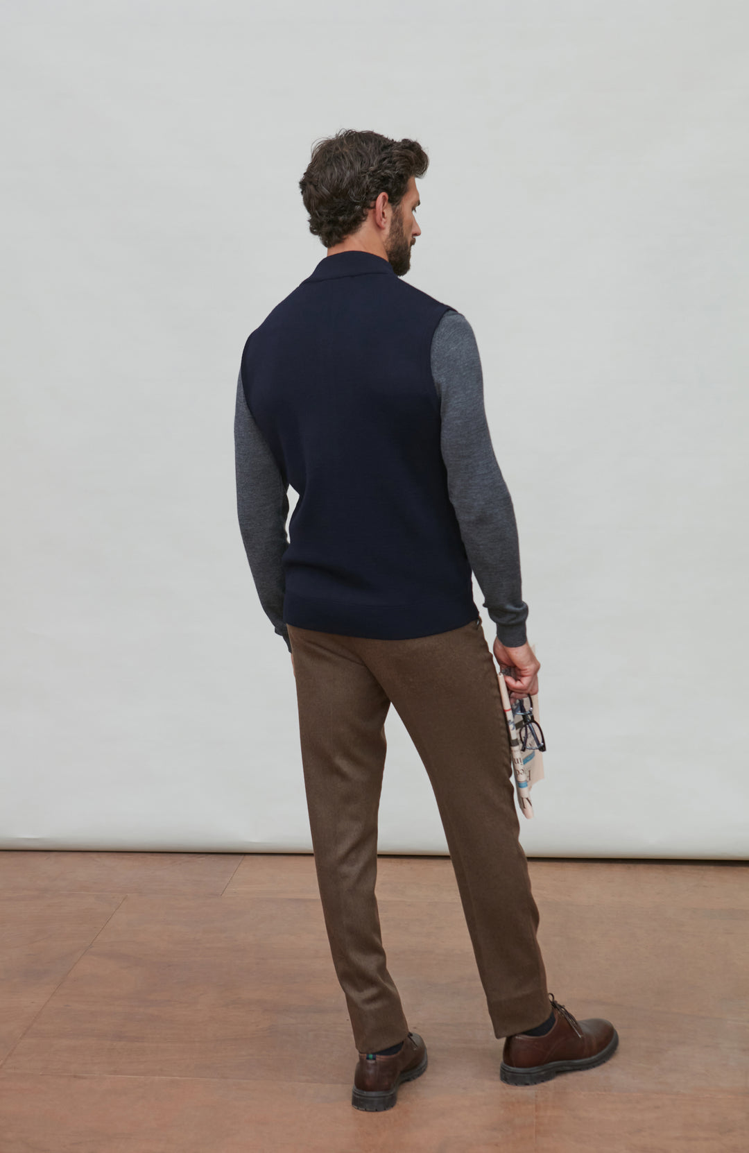 A man is shown from the back wearing a color-block knit sweater vest with navy blue and heather gray sleeves, paired with brown trousers and brown shoes, standing against a plain light gray wall.