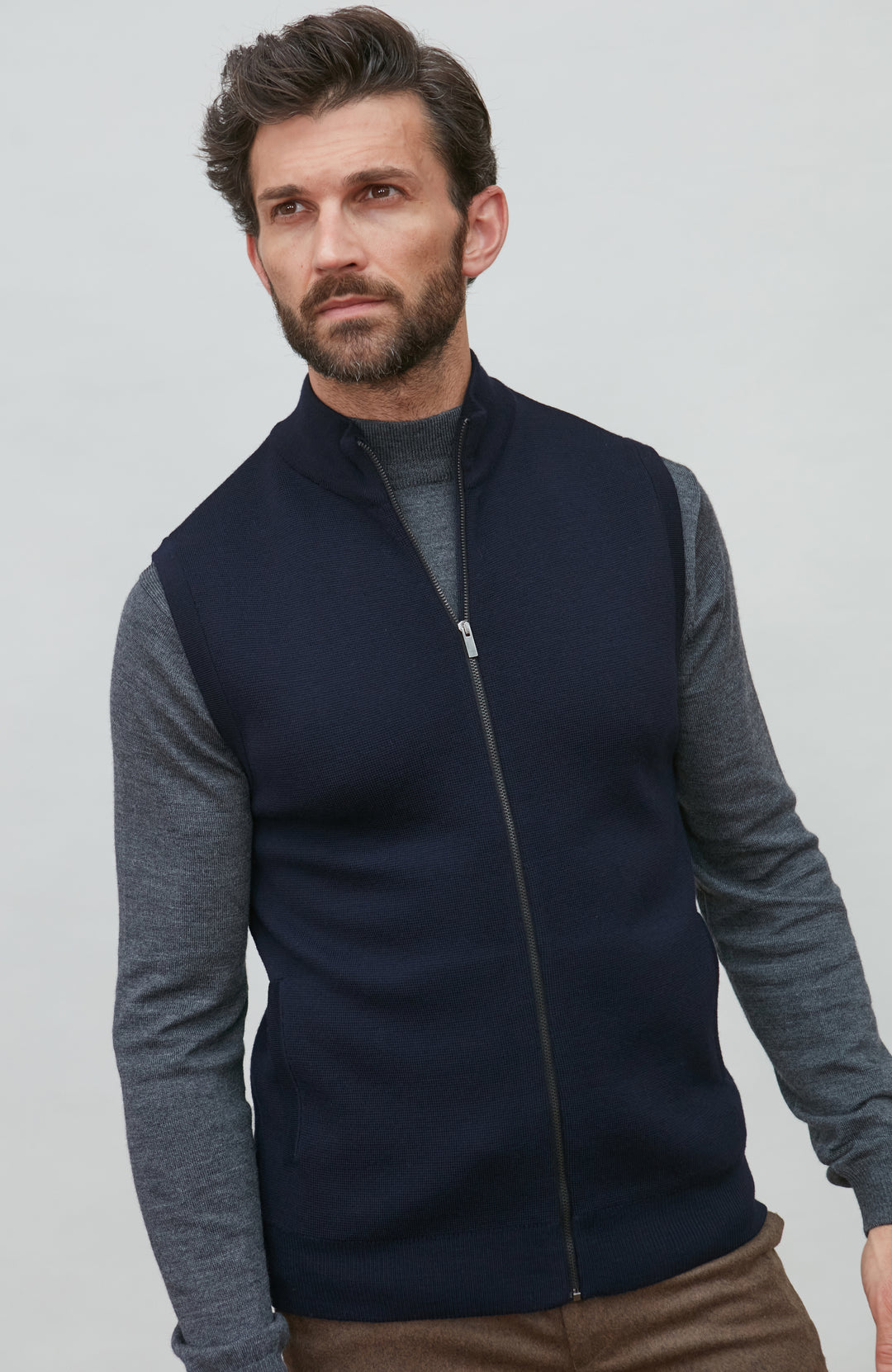 A man wearing a navy blue zip-up sleeveless merino wool vest by Paul James Knitwear over a gray long-sleeve shirt and brown trousers, photographed against a light gray studio background.