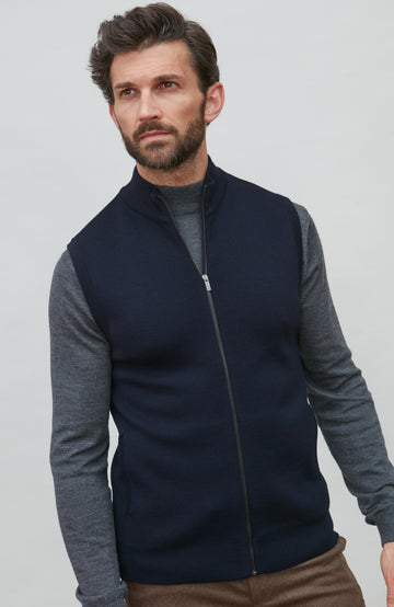 A man wearing a navy blue zip-up sleeveless merino wool vest by Paul James Knitwear over a gray long-sleeve shirt and brown trousers, photographed against a light gray studio background.