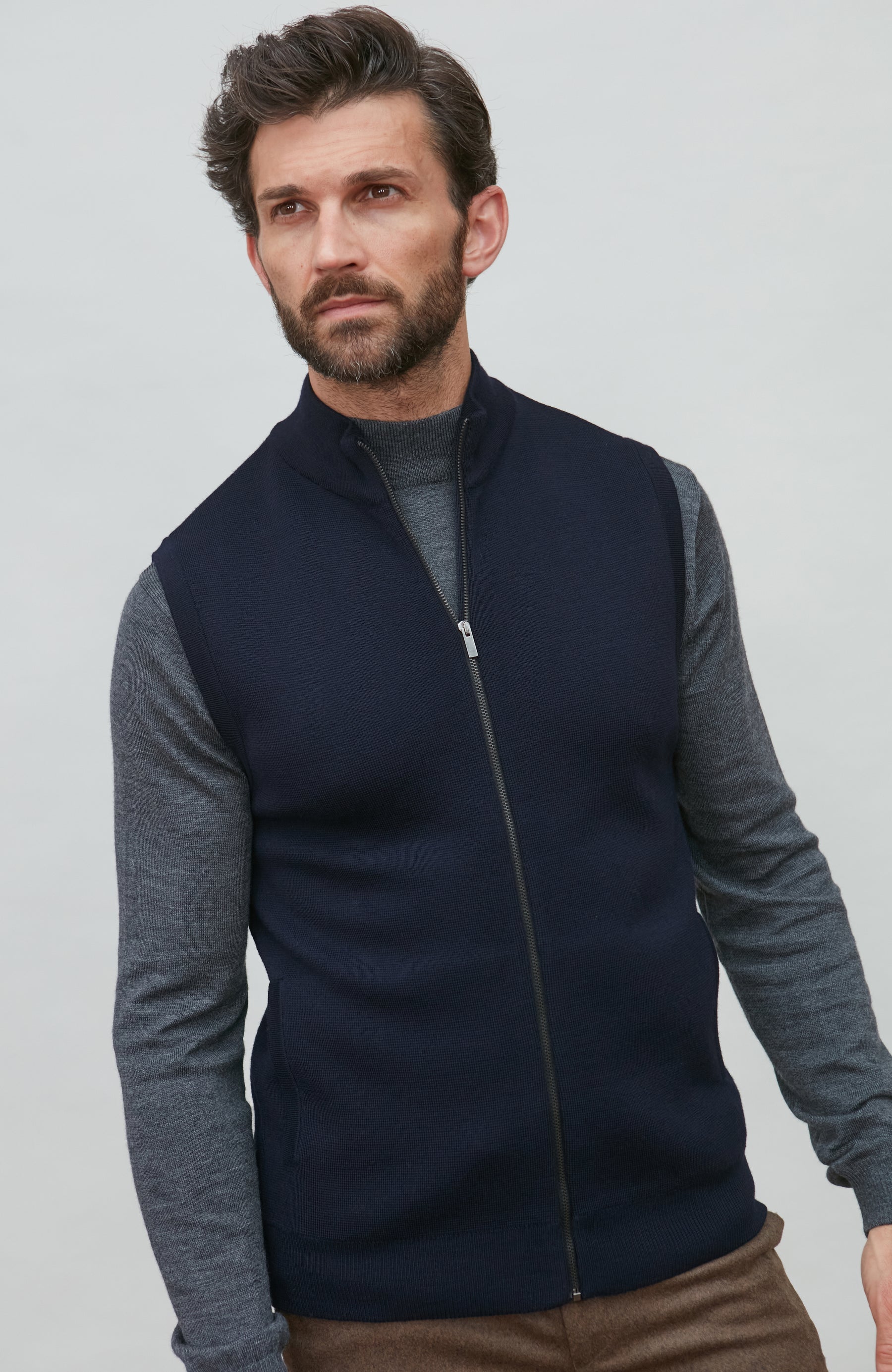 A man wearing a navy blue zip-up sleeveless merino wool vest by Paul James Knitwear over a gray long-sleeve shirt and brown trousers, photographed against a light gray studio background.