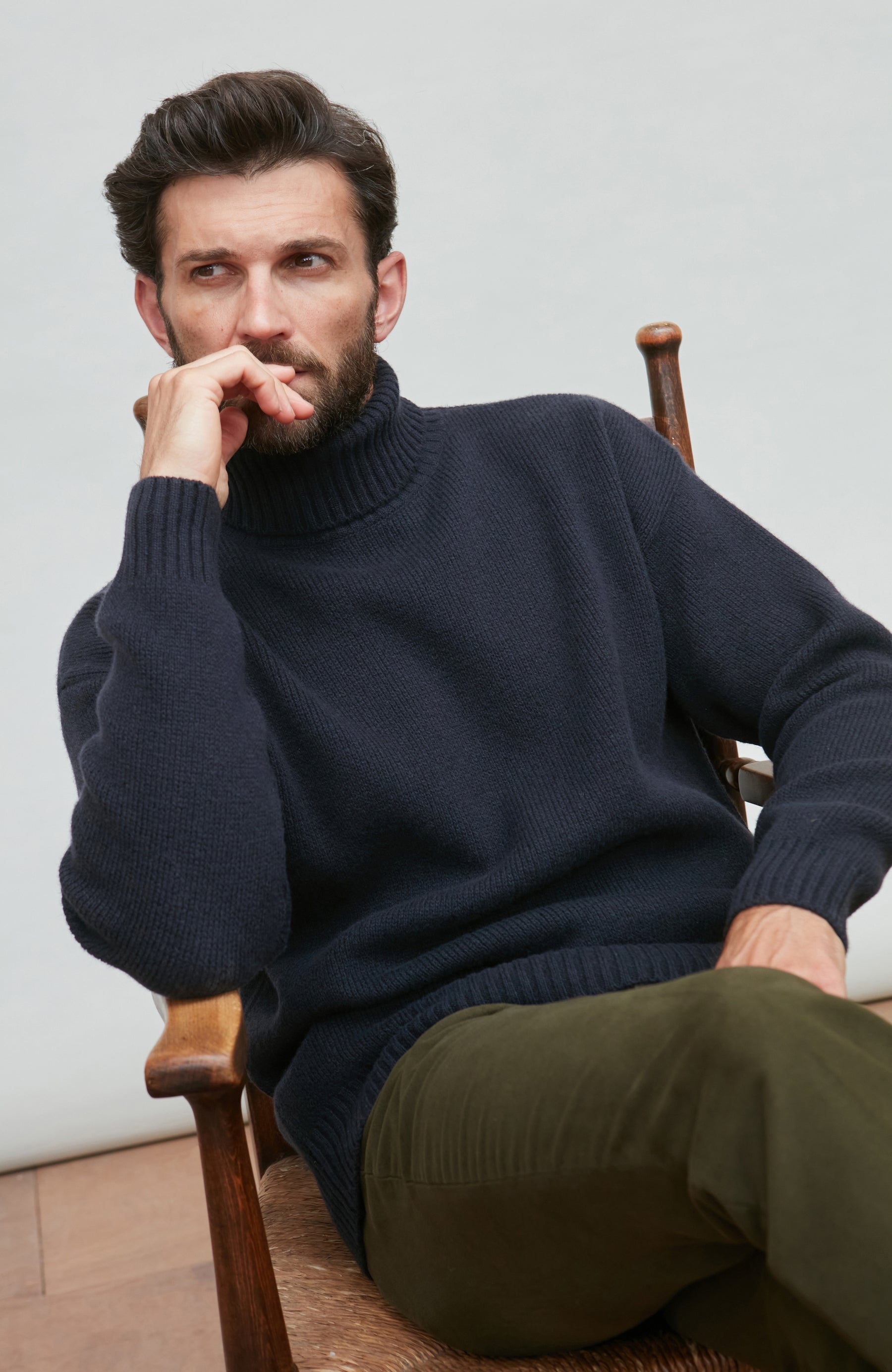 Man wearing a navy merino cashmere roll neck sweater and green pants sitting on a wooden chair.