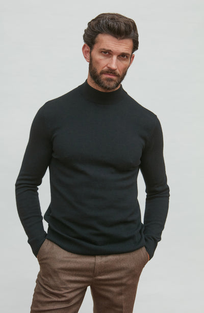 Man wearing a black merino cashmere turtleneck sweater and brown pants against a light gray background