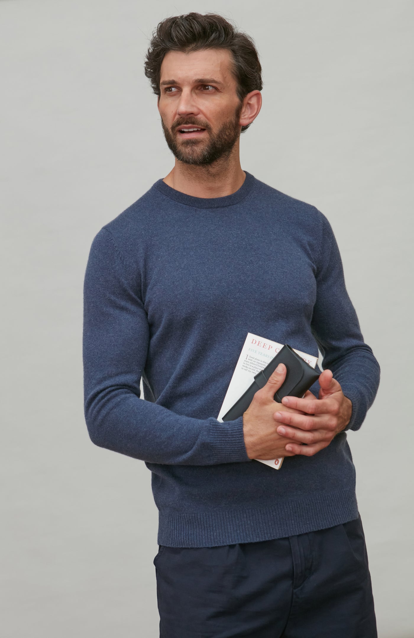 Man wearing a blue merino cashmere crew neck sweater holding a book against a gray background